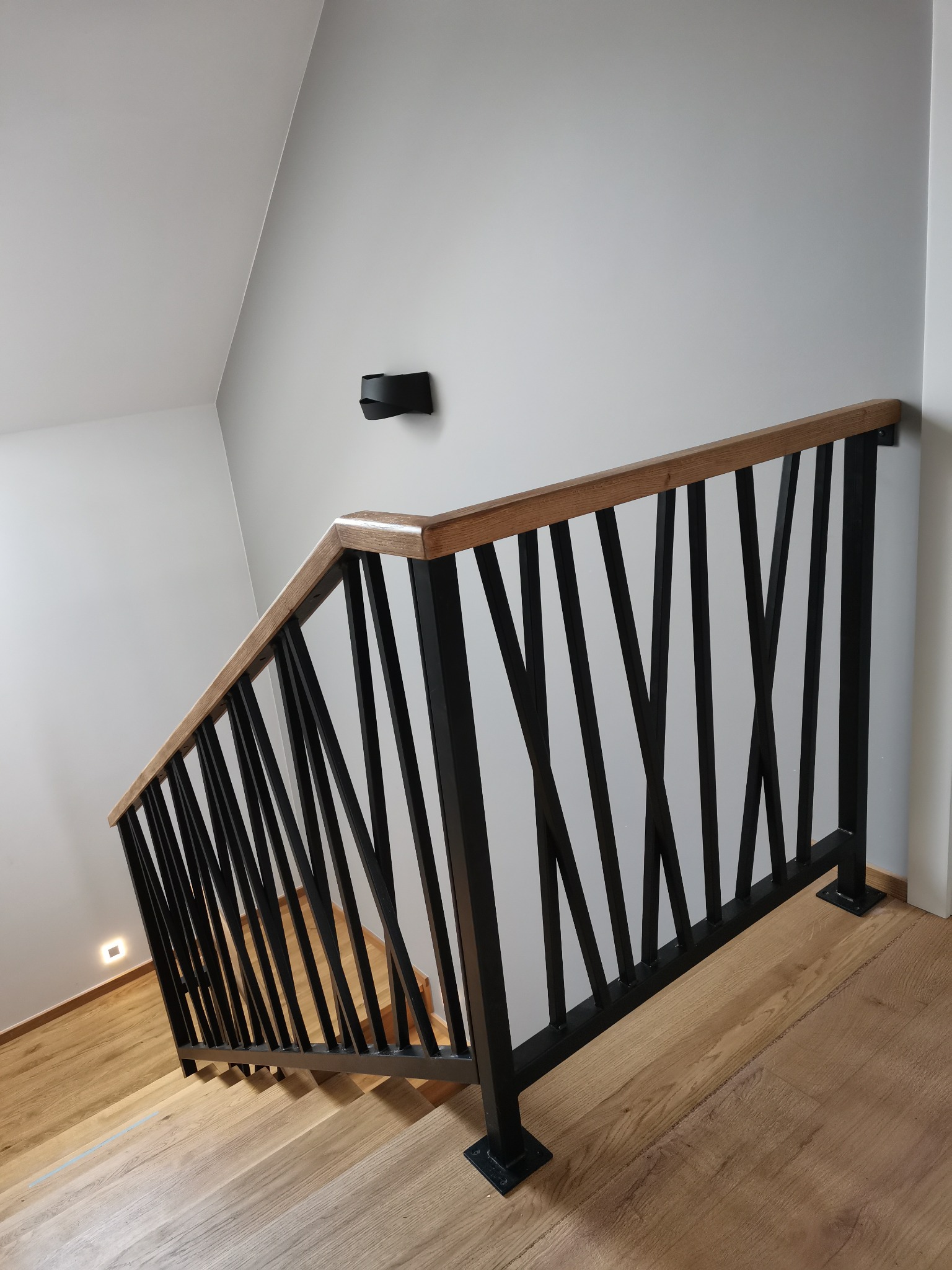 Nowoczesna balustrada schodowa z czarnymi, ukośnymi prętami i drewnianym poręczą, oświetlona przez minimalistyczny kinkiet na szarej ścianie.