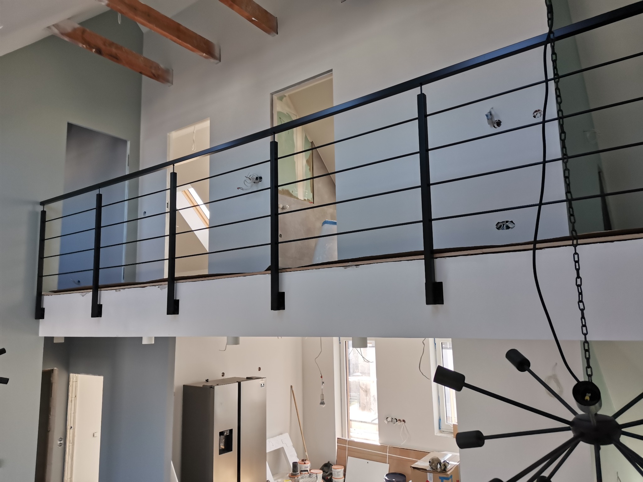 Czarna, pozioma barierka z cienkich prętów na antresoli w trakcie remontu, widoczne wystające kable i niedokończone ściany.