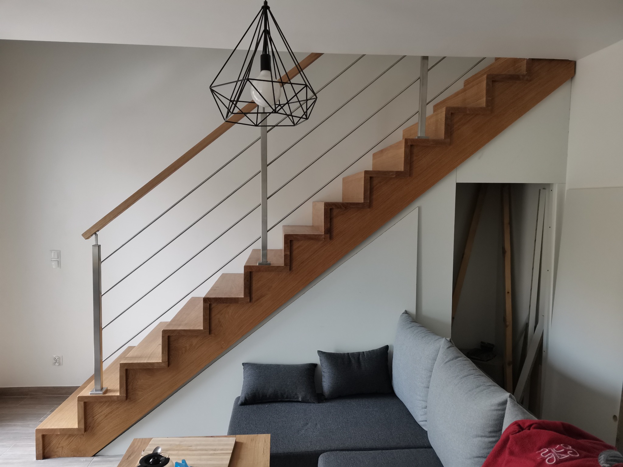 Drewniane schody z poziomymi, metalowymi barierkami w nowoczesnym wnętrzu z geometryczną lampą wiszącą.