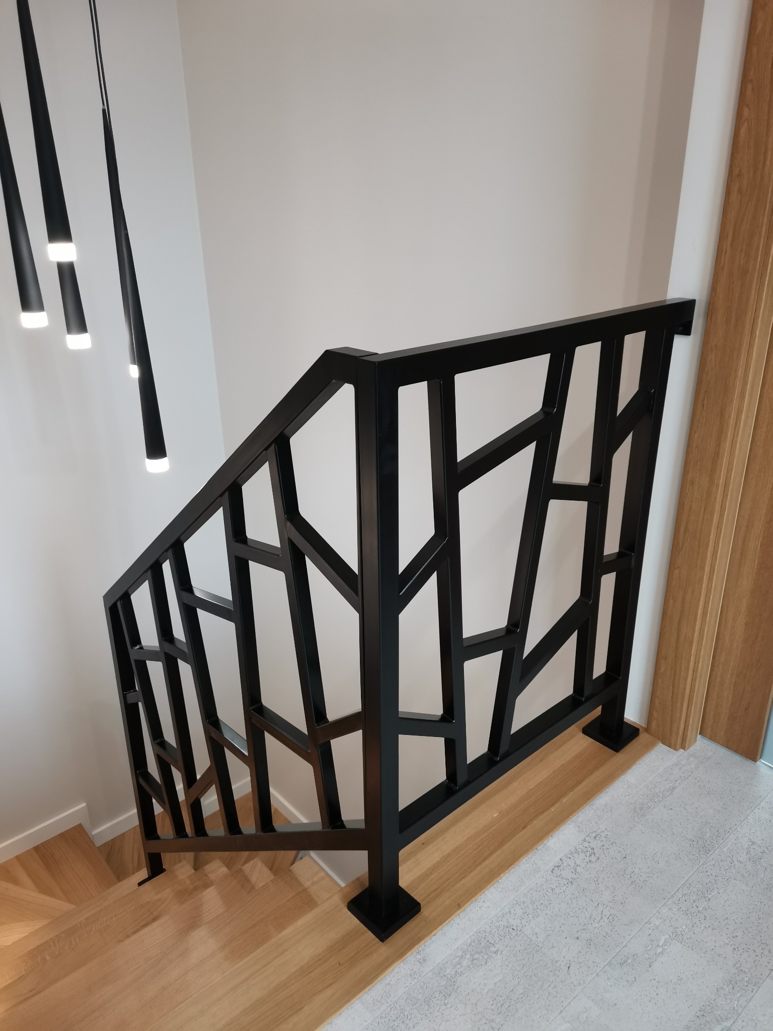 Nowoczesna, czarna balustrada schodowa z geometrycznym wzorem, zamontowana na drewnianych schodach, oświetlona wiszącymi lampami.