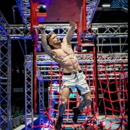 Mężczyzna o wysportowanej sylwetce, z napiętymi mięśniami brzucha, w trakcie pokonywania toru przeszkód Ninja Warrior, wisząc na rękach na metalowej konstrukcji z czerwonymi elementami, w tle...