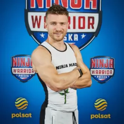 Uśmiechnięty mężczyzna w sportowym stroju z logo Ninja Warrior, pozujący na tle logotypów programu.