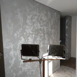 Ściana z dekoracyjną fakturą w odcieniach szarości oświetlona dwoma halogenami na statywie, widoczne ślady prac wykończeniowych na podłodze.