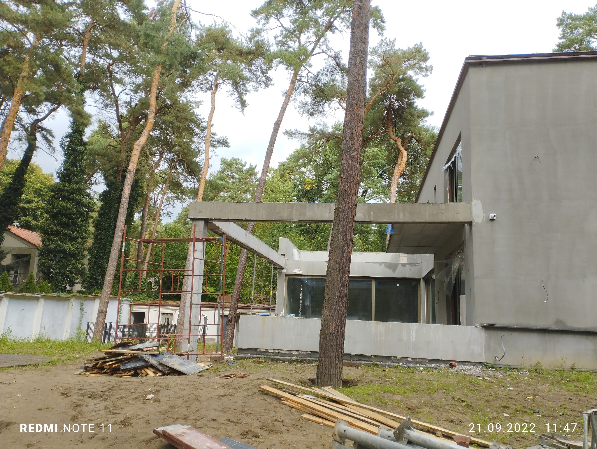 Surowy stan budynku z szarą elewacją w trakcie budowy, widoczne rusztowania, stos desek i wysokie drzewa w tle.