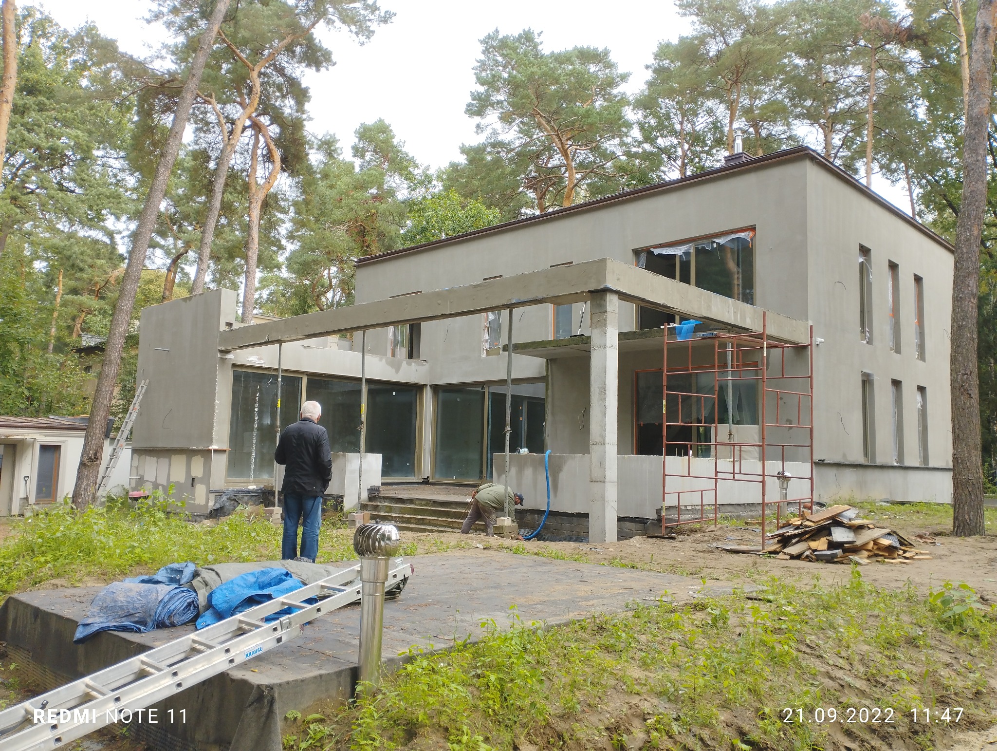 Dwukondygnacyjny dom w surowym stanie, z widocznymi rusztowaniami, przygotowany do dalszych prac wykończeniowych, z dwoma pracownikami na zewnątrz i stertą drewnianych desek obok