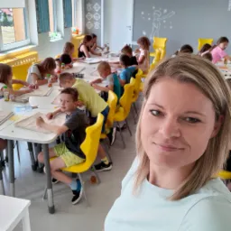 Kobieta robiąca selfie na tle dzieci uczestniczących w zajęciach plastycznych przy białych stołach i żółtych krzesłach w jasnym pomieszczeniu z dekoracjami na ścianach.