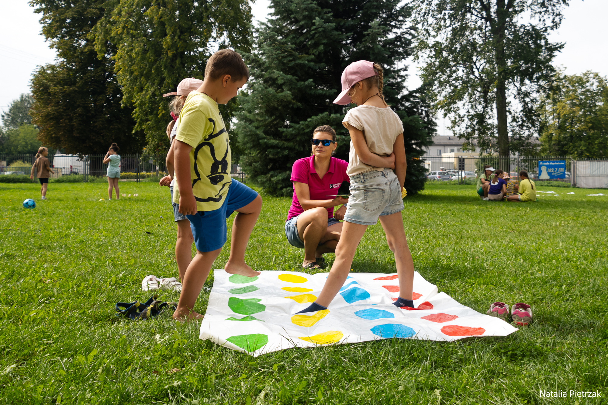 Dzieci grają w Twistera na trawie pod okiem kobiety w okularach słonecznych, w tle widoczne inne dzieci i drzewa.