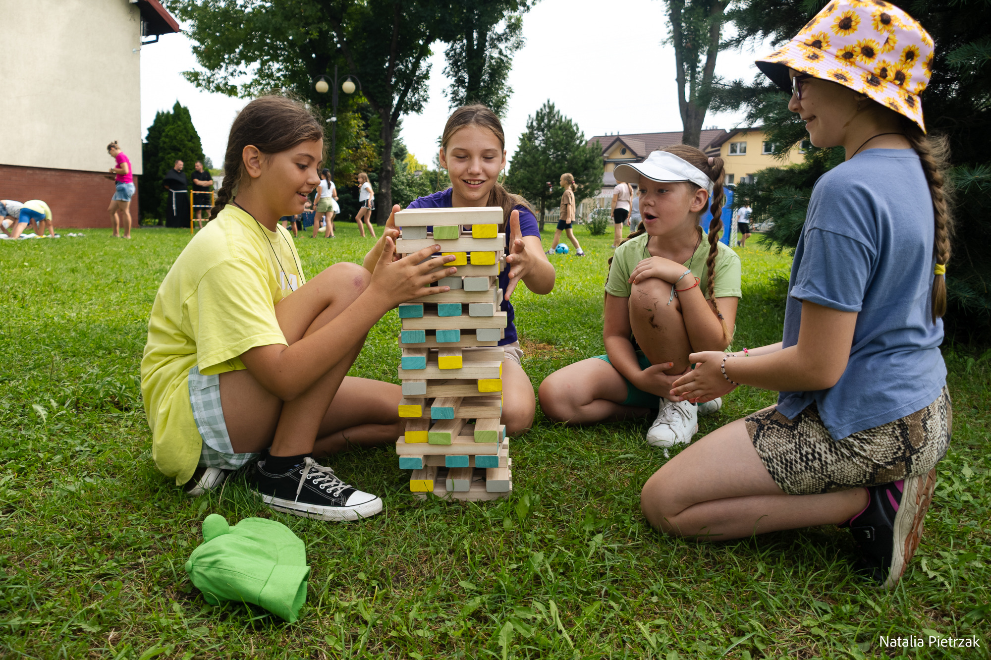 Cztery dziewczynki siedzą na trawie i grają w Jenga XXL; w tle widać inne dzieci i budynek.