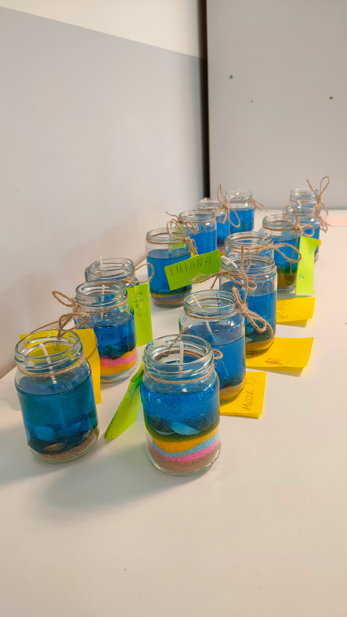 Rząd słoiczków z kolorowym piaskiem i niebieskim żelem, tworzących świeczki dekoracyjne, przewiązanych sznurkiem jutowym i oznaczonych karteczkami z imionami.