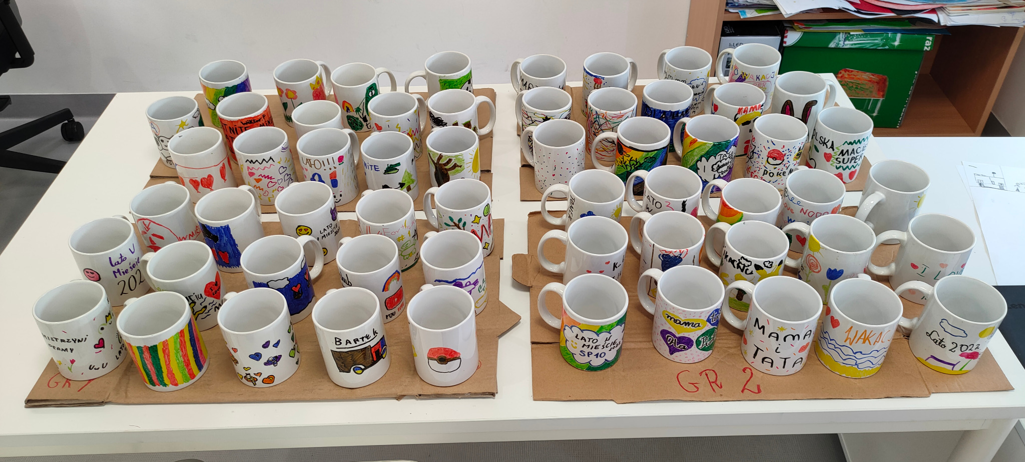 Grupa białych kubków, każdy z unikalnymi, kolorowymi rysunkami wykonanymi przez dzieci, ustawionych na tekturowych podkładkach na białym stole.