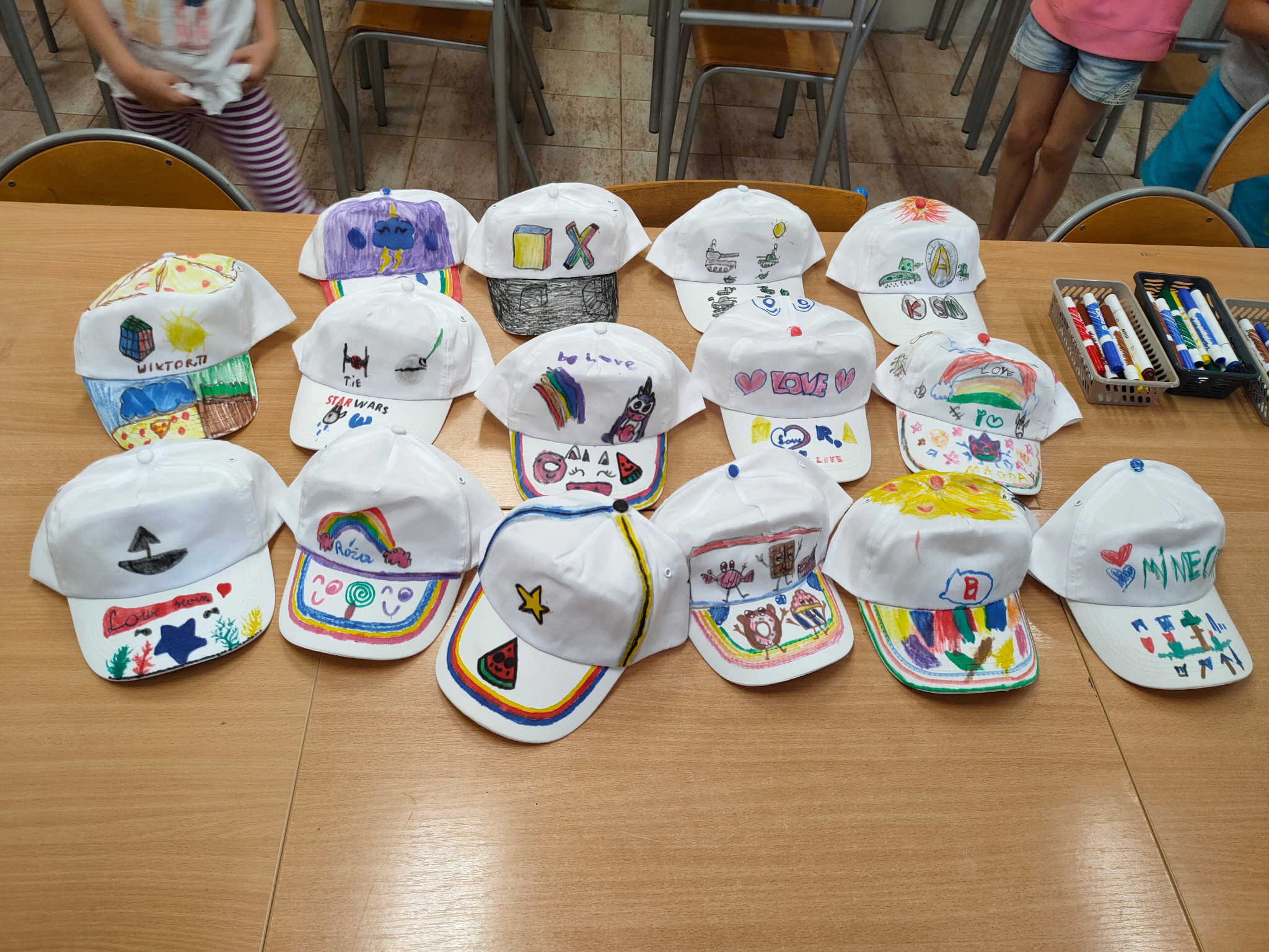 Warsztat artystyczny dla dzieci: Rząd białych czapek z daszkiem, każda ozdobiona unikalnymi, kolorowymi rysunkami wykonanymi przez dzieci, umieszczonych na drewnianym stole, z widocznymi pisakami...