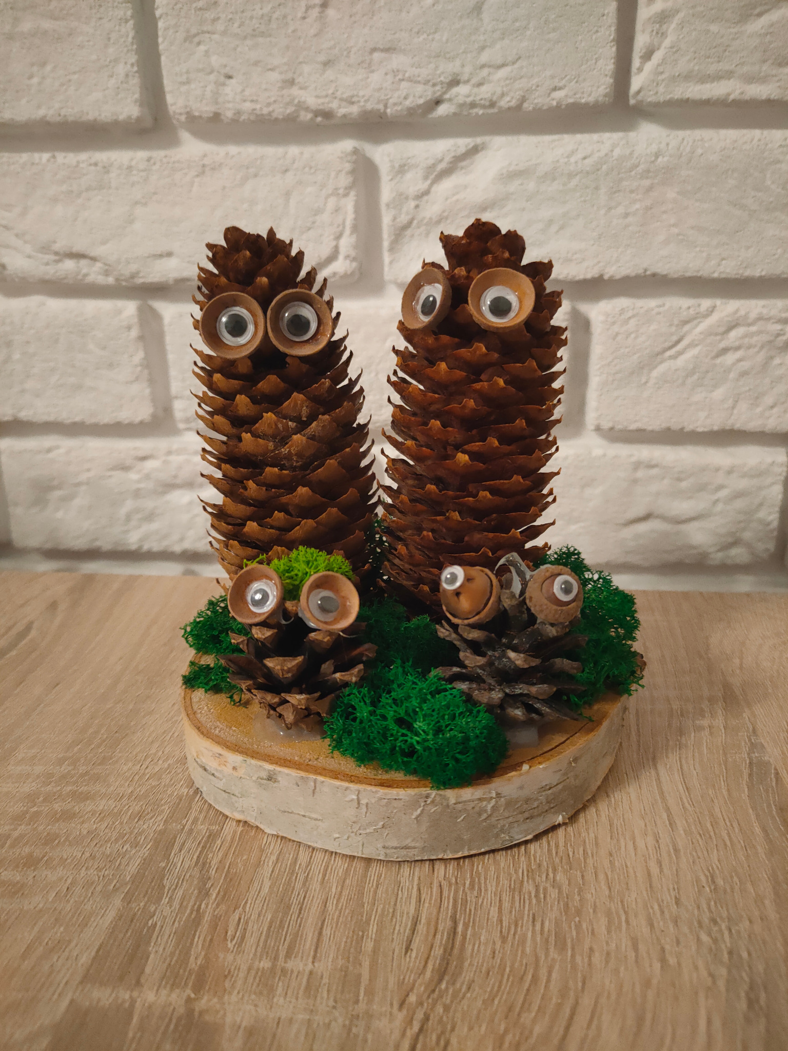 Dekoracja z szyszek i żołędzi z googly oczami, umieszczona na drewnianym krążku z mchem, na tle białej cegły.
