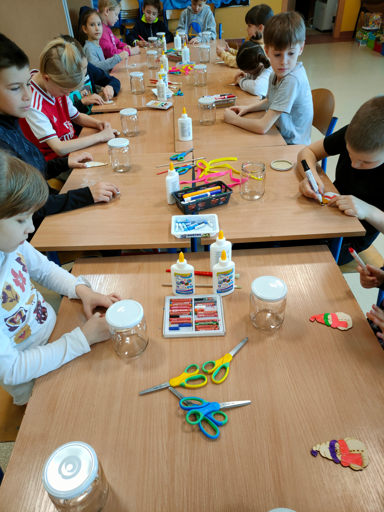 Dzieci przy drewnianych stołach, ozdabiają małe drewniane elementy i słoiki klejem, kredkami i nożyczkami, przygotowując dekoracje świąteczne.