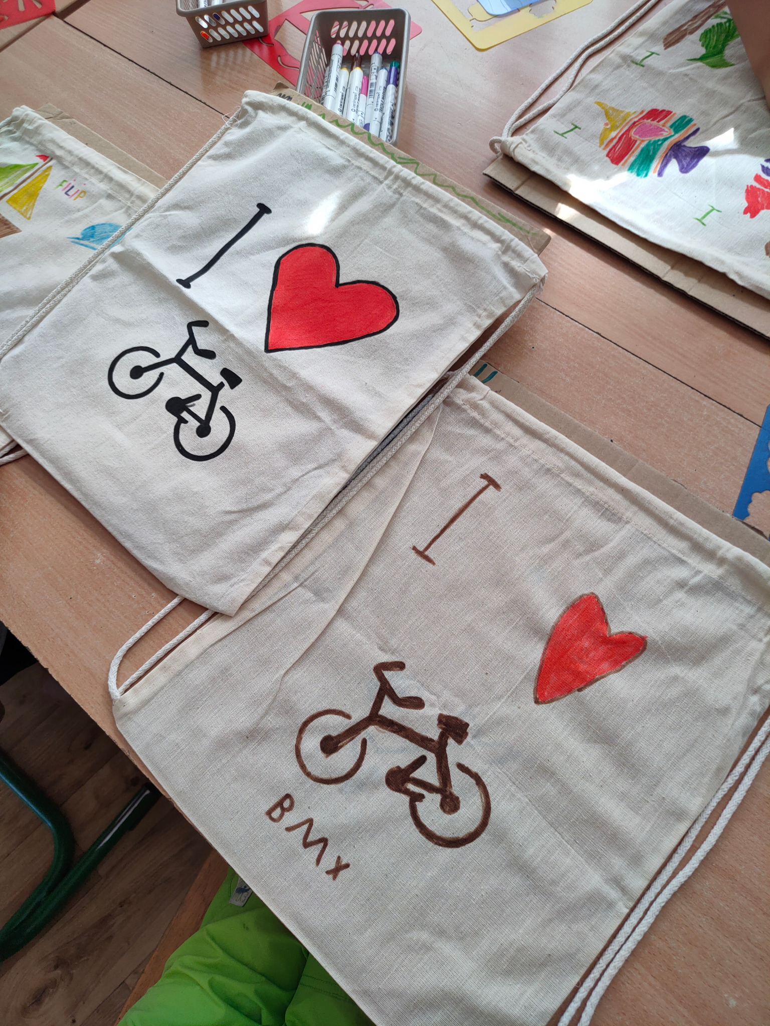 Warsztat plastyczny dla dzieci: ręcznie zdobione, płócienne worki z motywem roweru i serca, leżące na drewnianym stole.