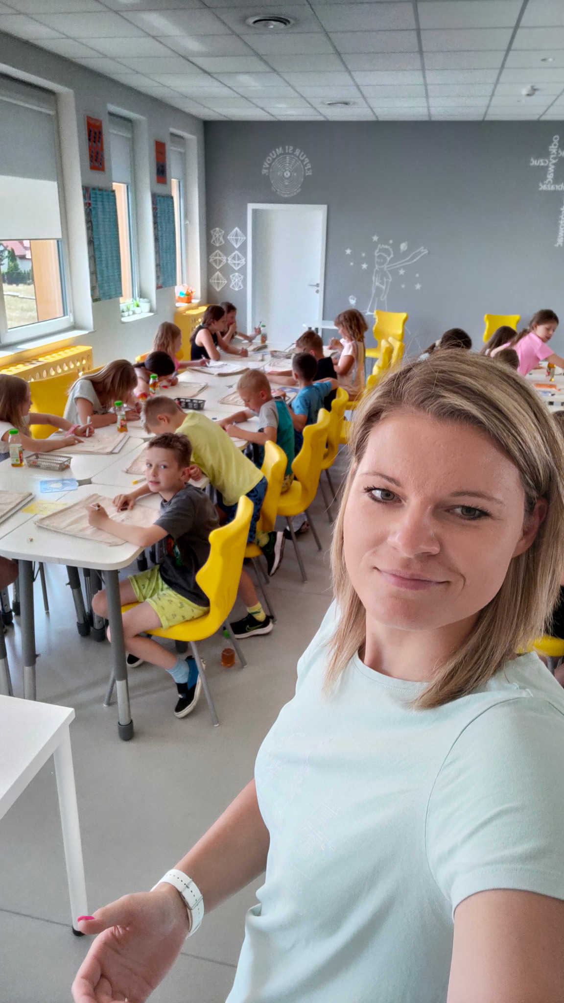 Kobieta robiąca selfie na tle dzieci uczestniczących w zajęciach plastycznych przy białych stołach i żółtych krzesłach w jasnym pomieszczeniu z dekoracjami na ścianach.