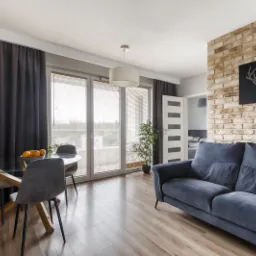 Jasne wnętrze salonu z ceglaną ścianą, szarą sofą, szklanym stołem z owocami i dużymi oknami z żaluzjami.