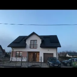 Nowoczesny dom jednorodzinny z ciemną dachówką i panelami słonecznymi na dachu, widoczny zza metalowego ogrodzenia, z zaparkowanym samochodem na podjeździe.