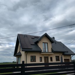 Przedsiębiorstwo Wielobranżowe Ms-Ortis Produkt Kobieracki, Selerowicz Sp. j. - Dom jednorodzinny z panelami fotowoltaicznymi na dachu, widoczny zza czarnego ogrodzenia, pochmurne niebo w tle.