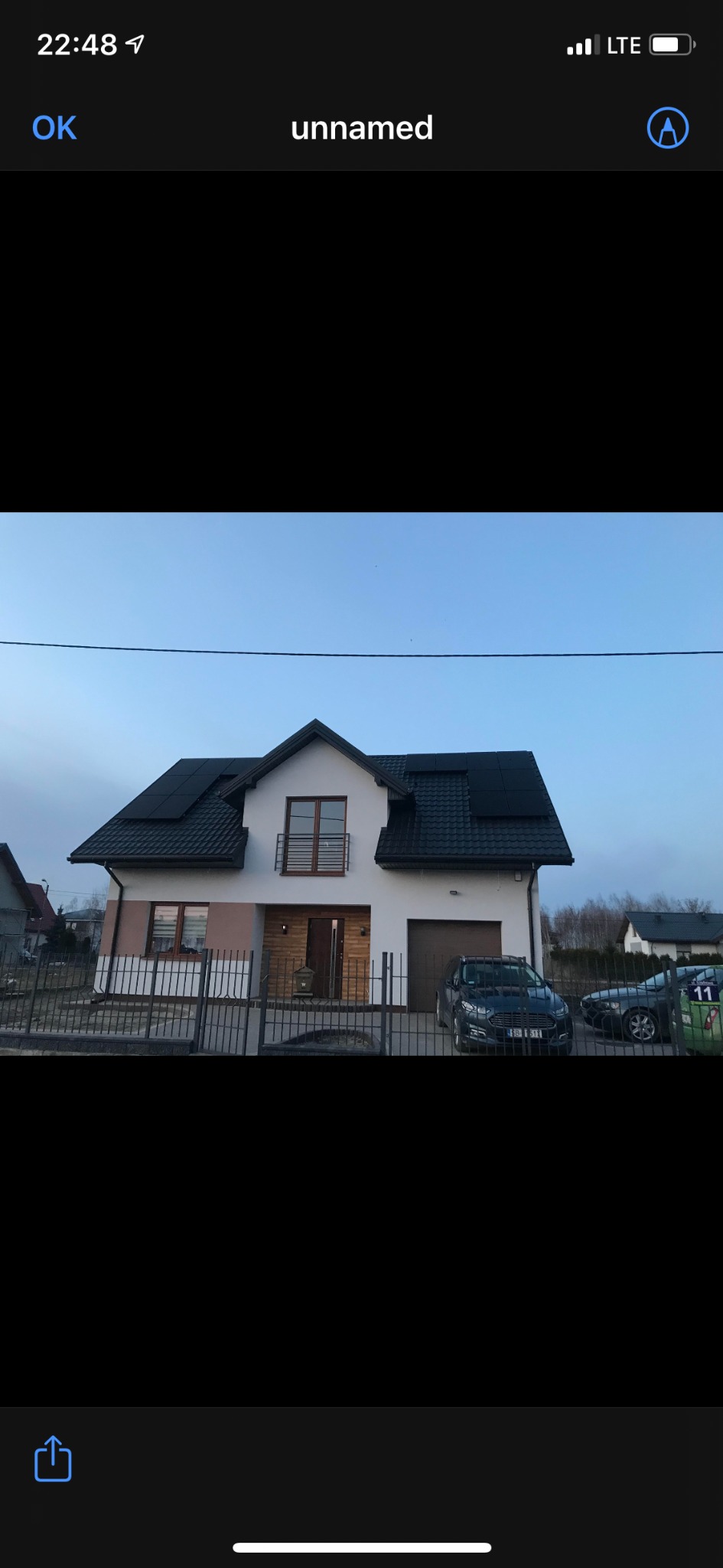 Nowoczesny dom jednorodzinny z ciemną dachówką i panelami słonecznymi na dachu, widoczny zza metalowego ogrodzenia, z zaparkowanym samochodem na podjeździe.