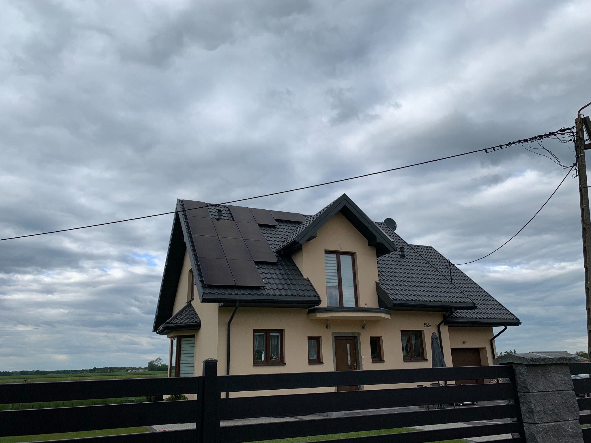 Dom jednorodzinny z panelami fotowoltaicznymi na dachu, widoczny zza czarnego ogrodzenia, pochmurne niebo w tle.