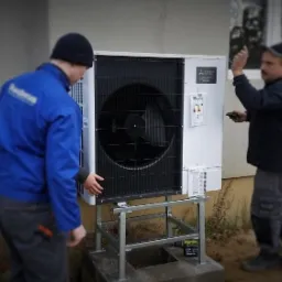 Dwóch instalatorów montuje zewnętrzną jednostkę pompy ciepła na metalowej konstrukcji przy ścianie budynku. Jeden instalator w niebieskiej kurtce dotyka urządzenia, drugi trzyma pilot.