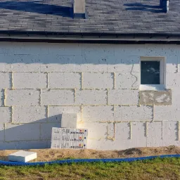 Ściana budynku z białych bloczków, częściowo wypełniona pianką montażową, z niebieską rurą wentylacyjną wzdłuż elewacji i paczkami styropianu Swissopor u podstawy ściany.