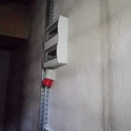 Biała skrzynka rozdzielcza z czerwonym gniazdem siłowym zamontowana na metalowej szynie perforowanej na szarej, nierównej ścianie w pomieszczeniu technicznym.