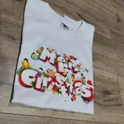Biała koszulka z nadrukiem 'Merry Christmas' w trójwymiarowym stylu, ozdobiona motywami świątecznymi, leżąca na drewnianym podłożu.