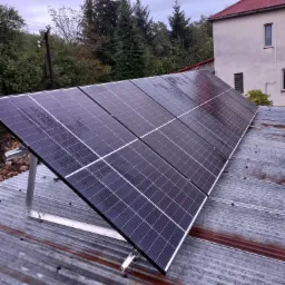 Panel słoneczny zainstalowany na blaszanym dachu budynku mieszkalnego, widoczne drzewa i pochmurne niebo w tle, metalowa konstrukcja wsporcza.