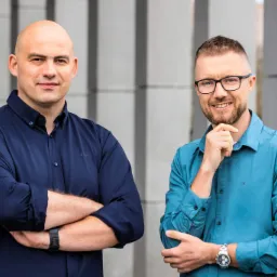 Portret dwóch mężczyzn w koszulach na tle nowoczesnej fasady budynku, uśmiechnięci i patrzący w obiektyw.