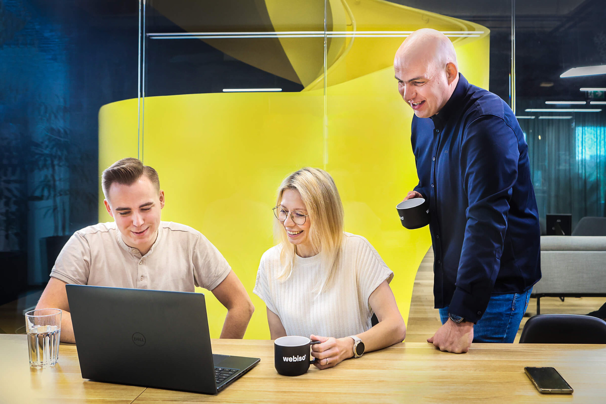 Trzy osoby w biurze, dwie siedzące przy laptopie Dell i uśmiechnięte, trzecia stojąca z kubkiem 'webiso' w ręku, w tle żółta konstrukcja.