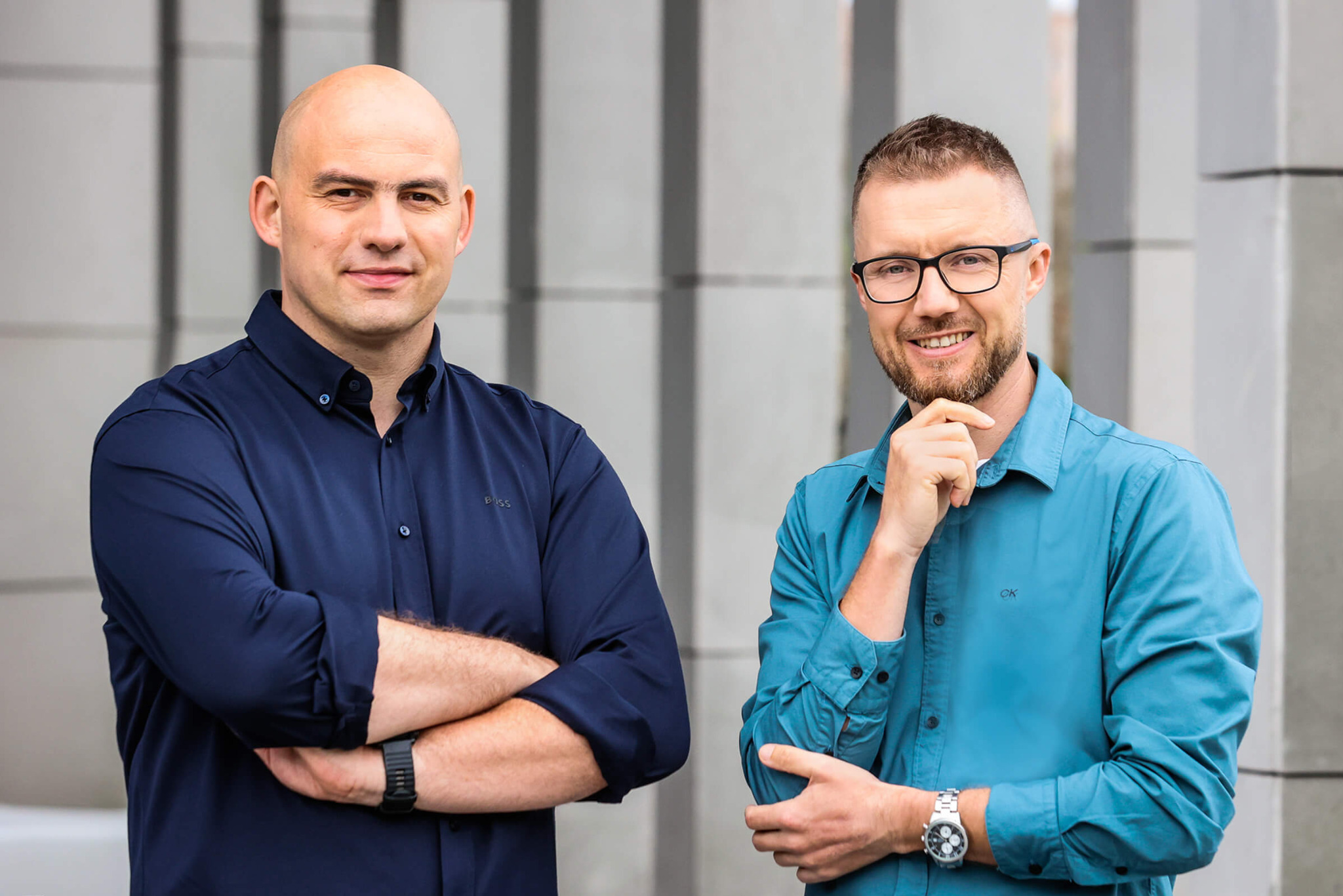 Portret dwóch mężczyzn w koszulach na tle nowoczesnej fasady budynku, uśmiechnięci i patrzący w obiektyw.