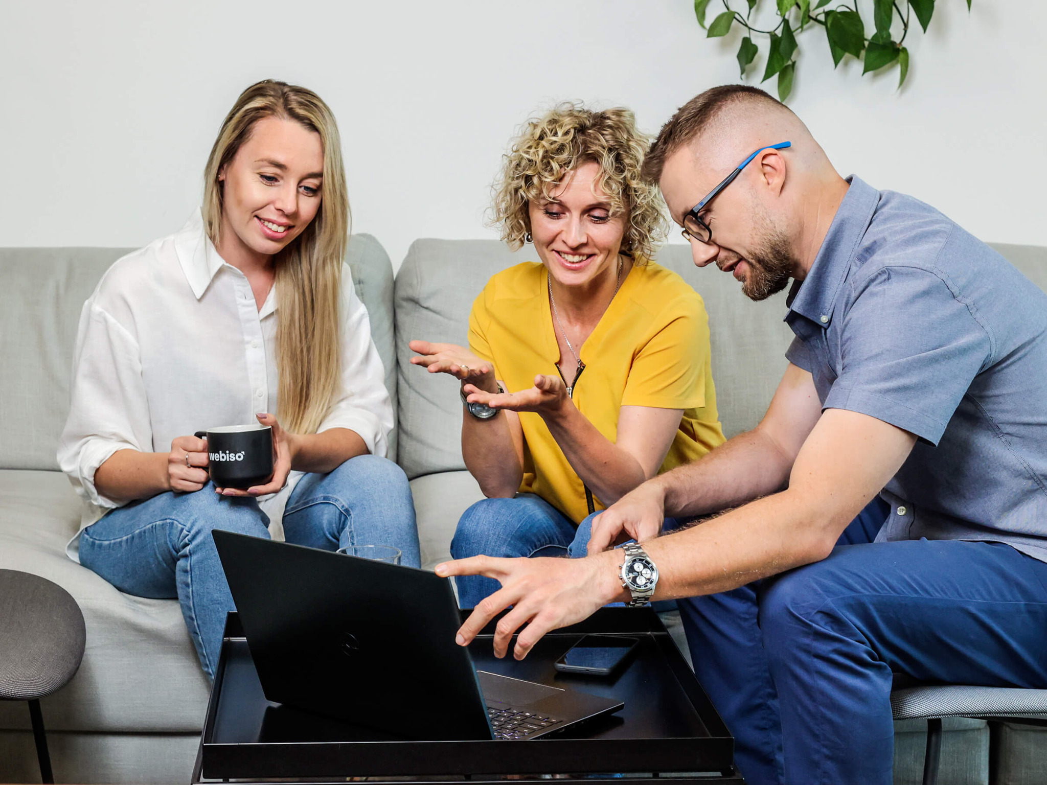Trzy osoby omawiają projekt na laptopie siedząc na kanapie, kobieta z blond włosami trzyma kubek z logo 'Webiso', roślina w tle.