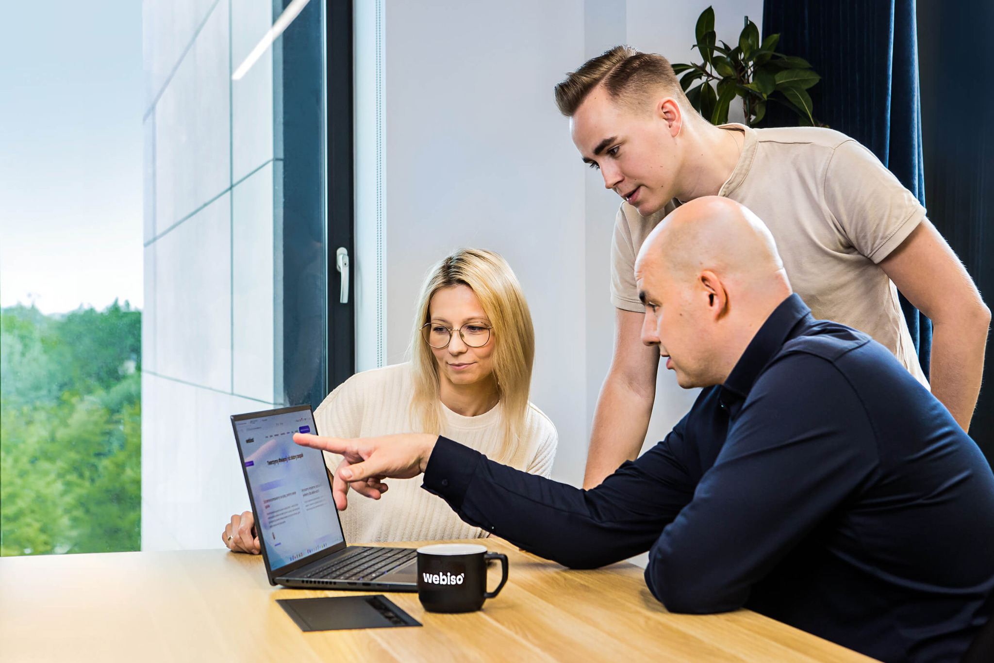 Zespół trzech osób analizuje zawartość strony internetowej wyświetlanej na laptopie, kubek z logo 'webiso' na stole.