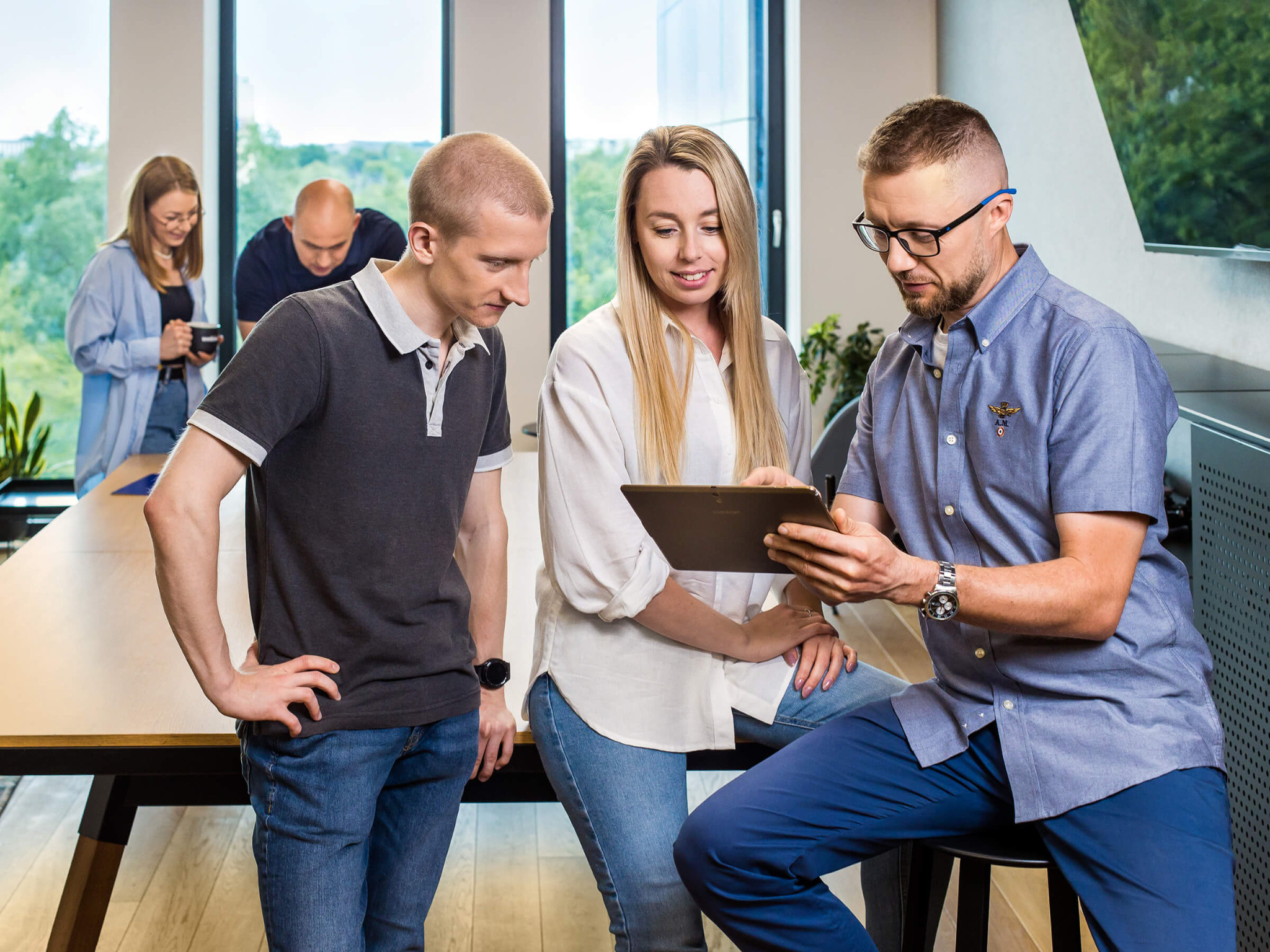 Zespół trzech osób analizuje zawartość tabletu w jasnym biurze; w tle dwie osoby przy stole, duże okna z zielenią na zewnątrz.