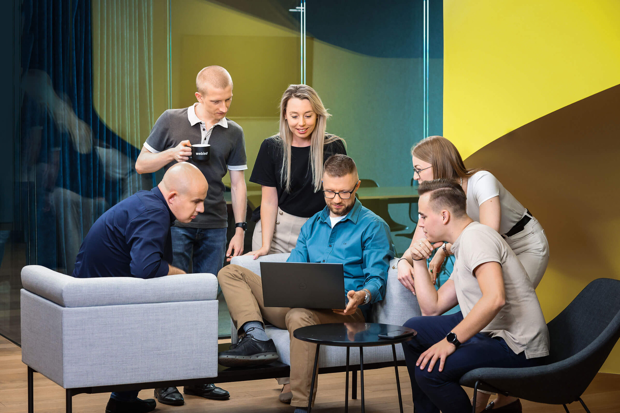 Zespół sześciu osób analizuje projekt strony internetowej na laptopie w nowoczesnym biurze, z kubkiem kawy w ręku jednego z pracowników.