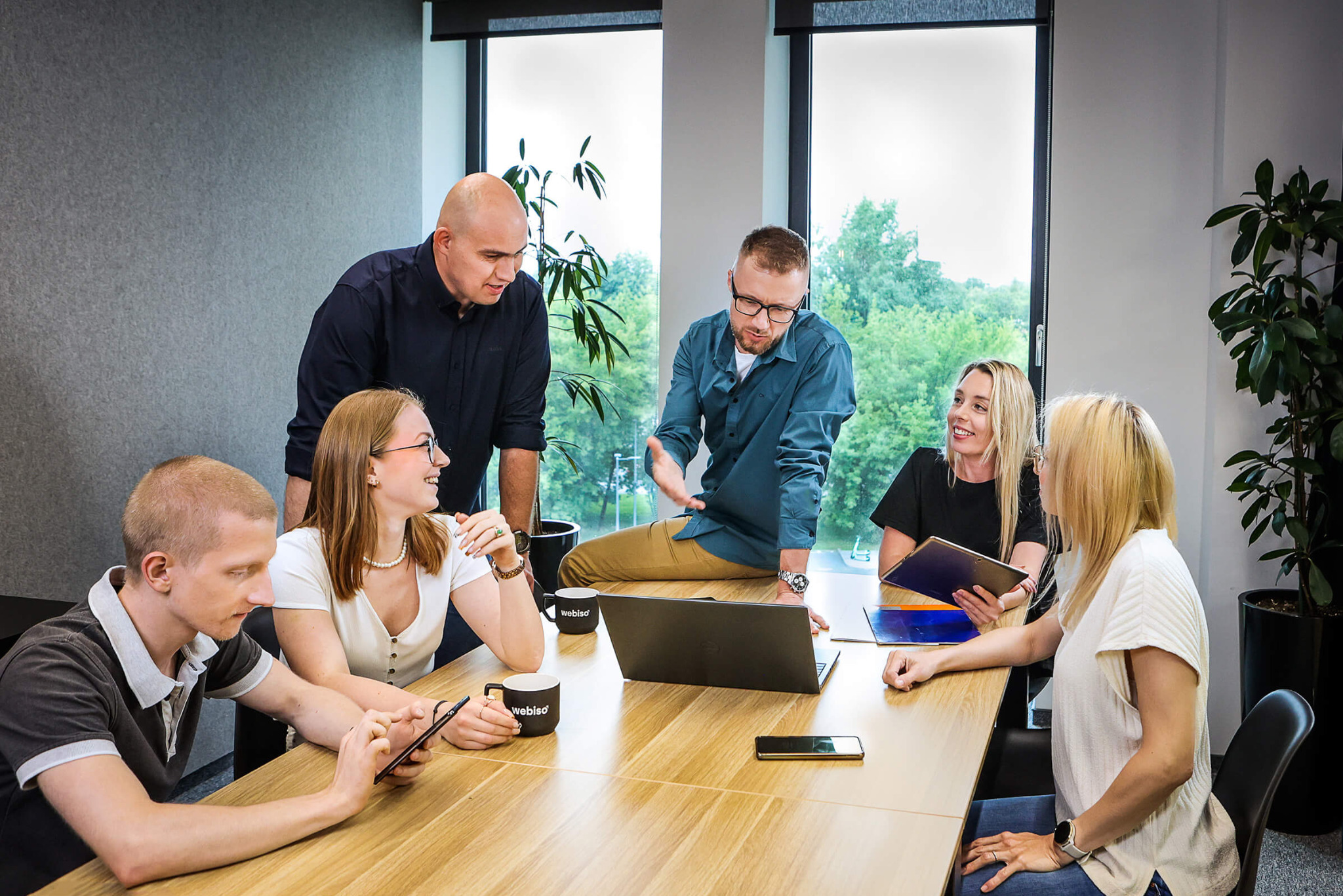 Zespół Webiso omawia strategię przy biurku, z laptopem i kubkami firmowymi, w tle widok za oknem.