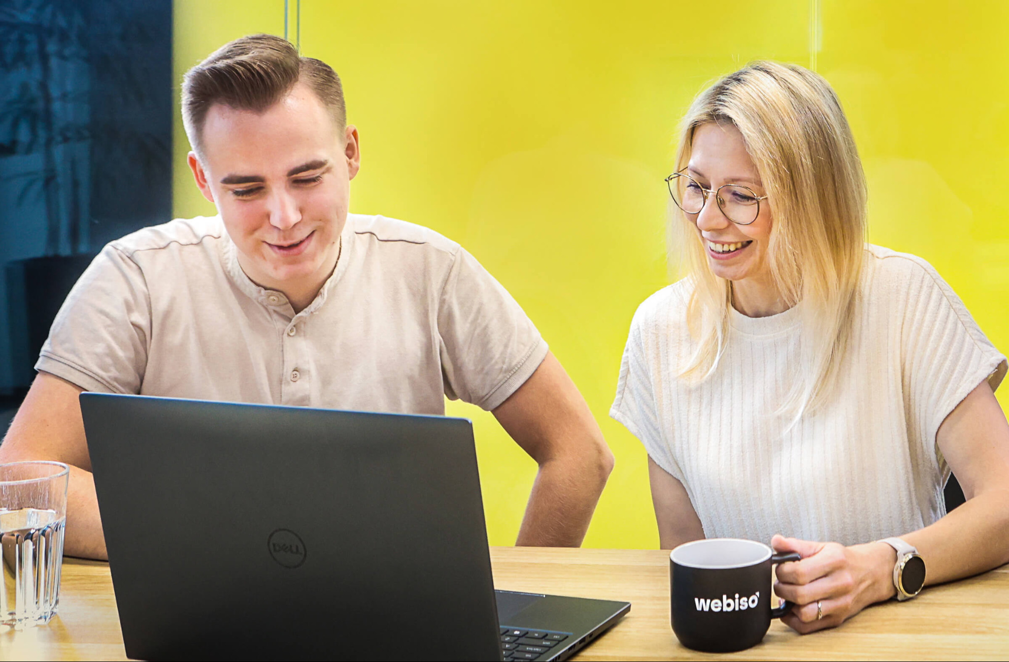Dwoje uśmiechniętych specjalistów, mężczyzna i kobieta w okularach, przegląda laptopa Dell na drewnianym biurku z kubkiem Webiso i szklanką wody na tle żółtej ściany w biurze.