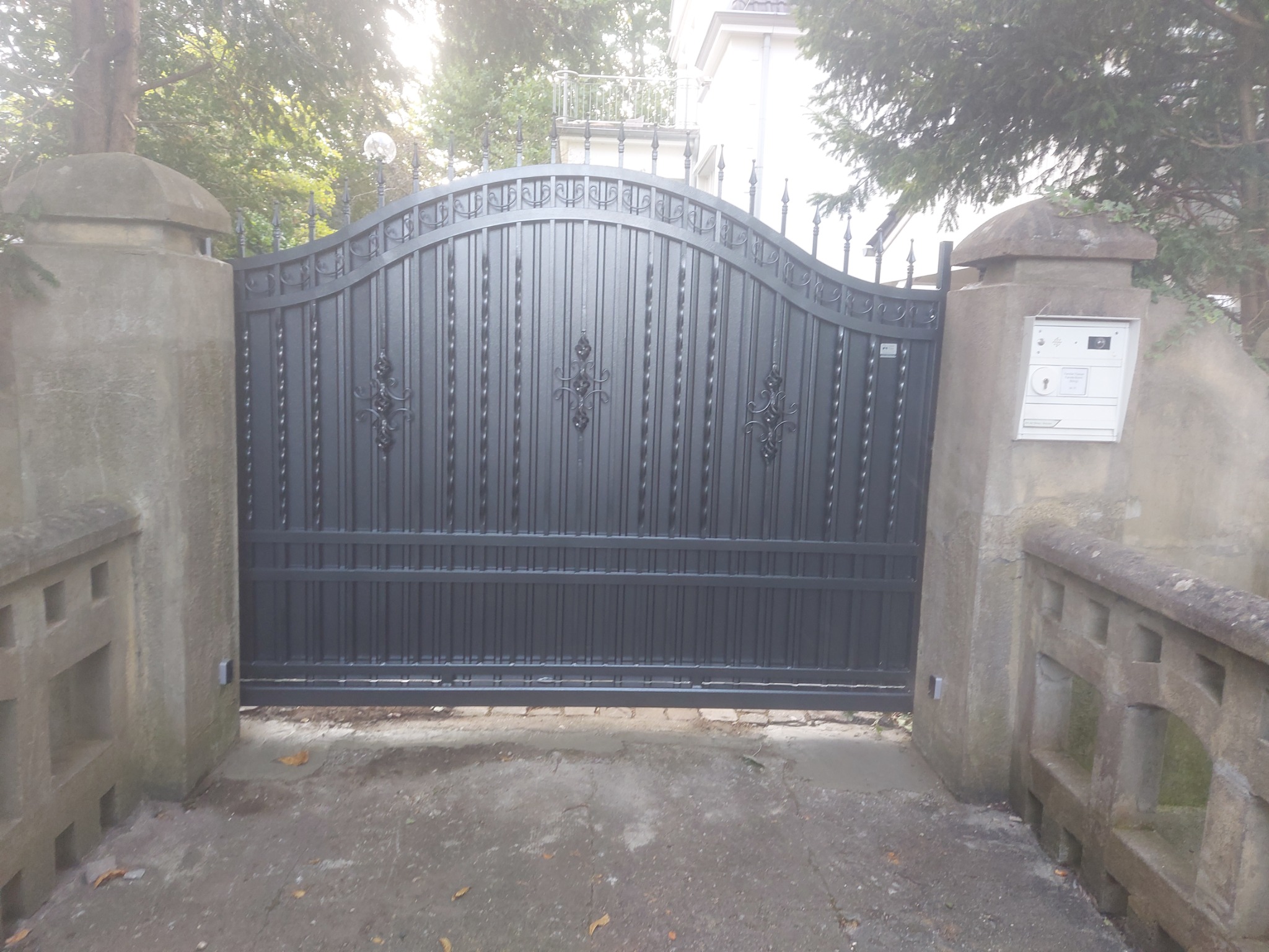 Czarna, metalowa brama wjazdowa z dekoracyjnymi elementami, osadzona pomiędzy betonowymi słupkami z zamontowaną skrzynką na listy, widok z zewnątrz posesji.
