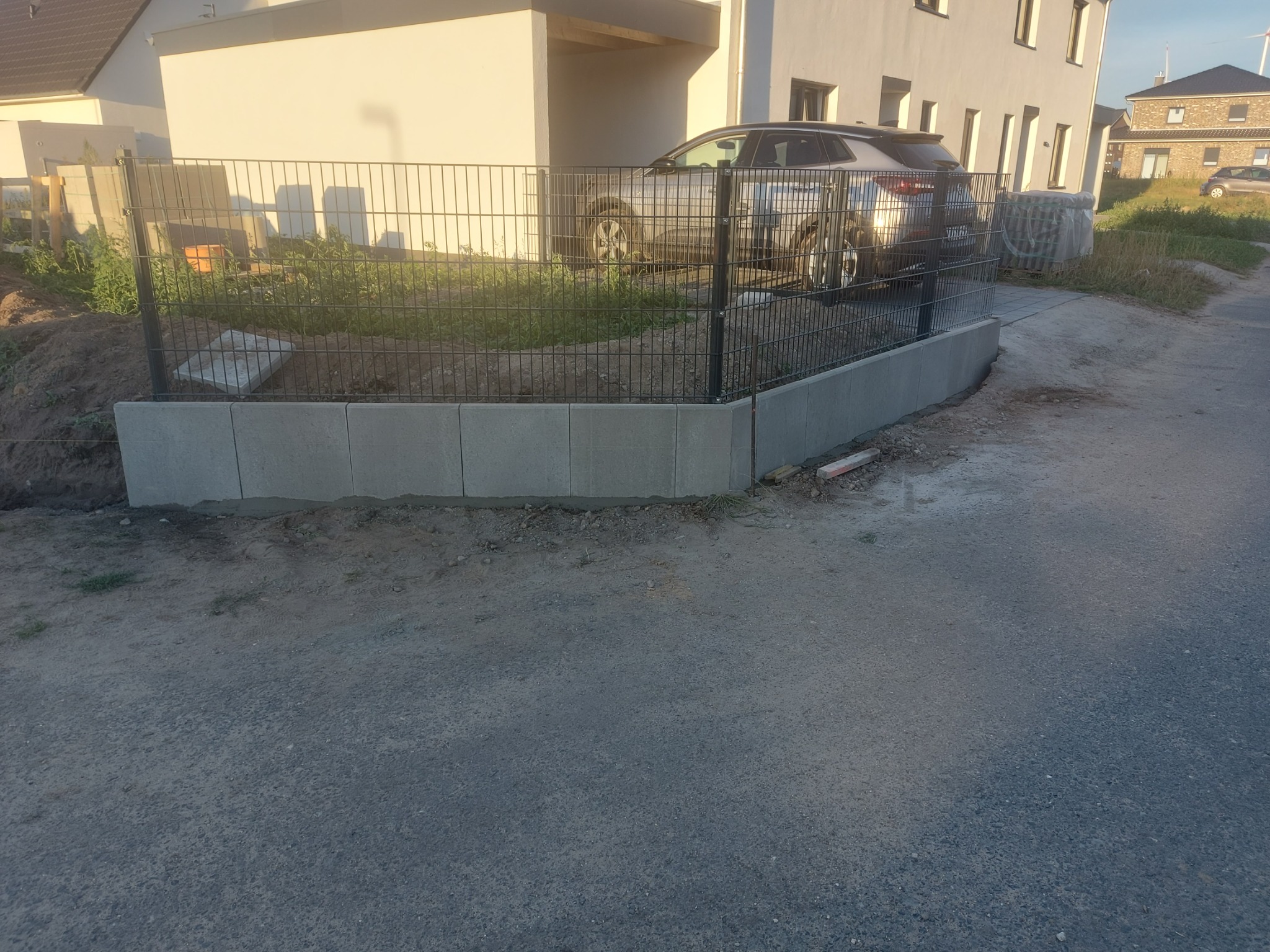 Ciemna metalowa siatka ogrodzeniowa na betonowej podmurówce, widok z zewnątrz posesji, samochód za ogrodzeniem, nowy dom w tle.