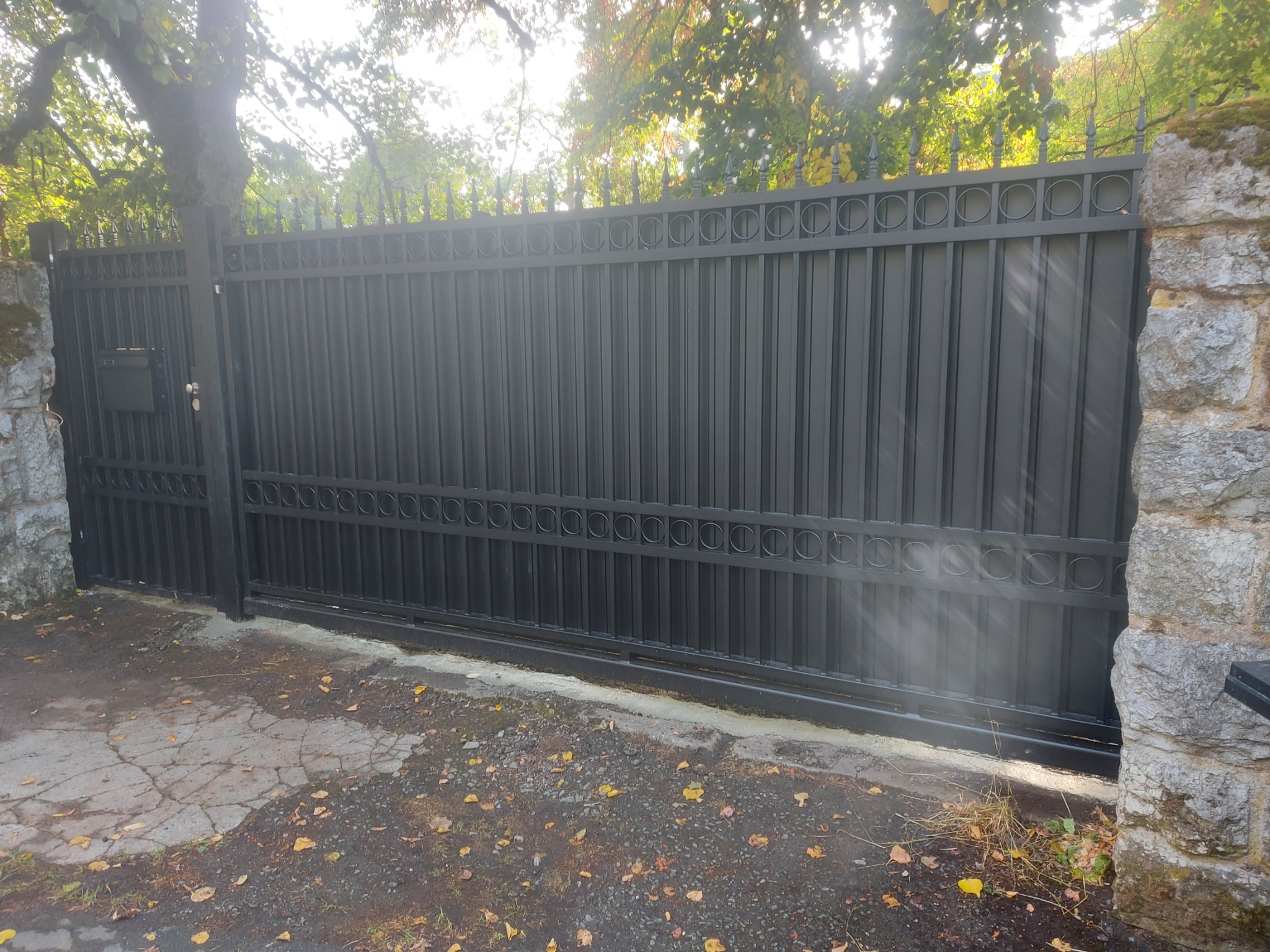 Nowoczesna, przesuwna brama w kolorze grafitowym z dekoracyjnymi okręgami u góry i dołu, zamontowana między kamiennymi słupkami, z widoczną skrzynką na listy i jesiennymi liśćmi na ziemi.