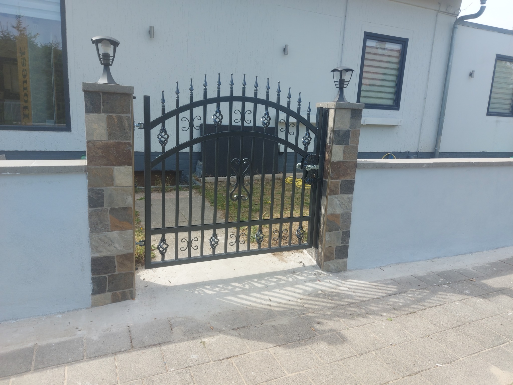 Kuta, czarna brama z dekoracyjnymi elementami, osadzona na murowanych słupkach z oświetleniem, na tle białego budynku z oknami.