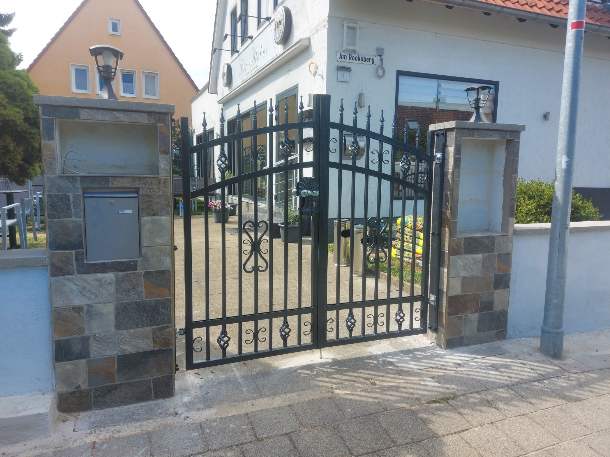 Otwarta, czarna, metalowa brama z dekoracyjnymi elementami, osadzona na kamiennych słupkach z wnękami i lampami, prowadząca na posesję z budynkiem o białej elewacji i szyldem 'Am Rooksberg'.