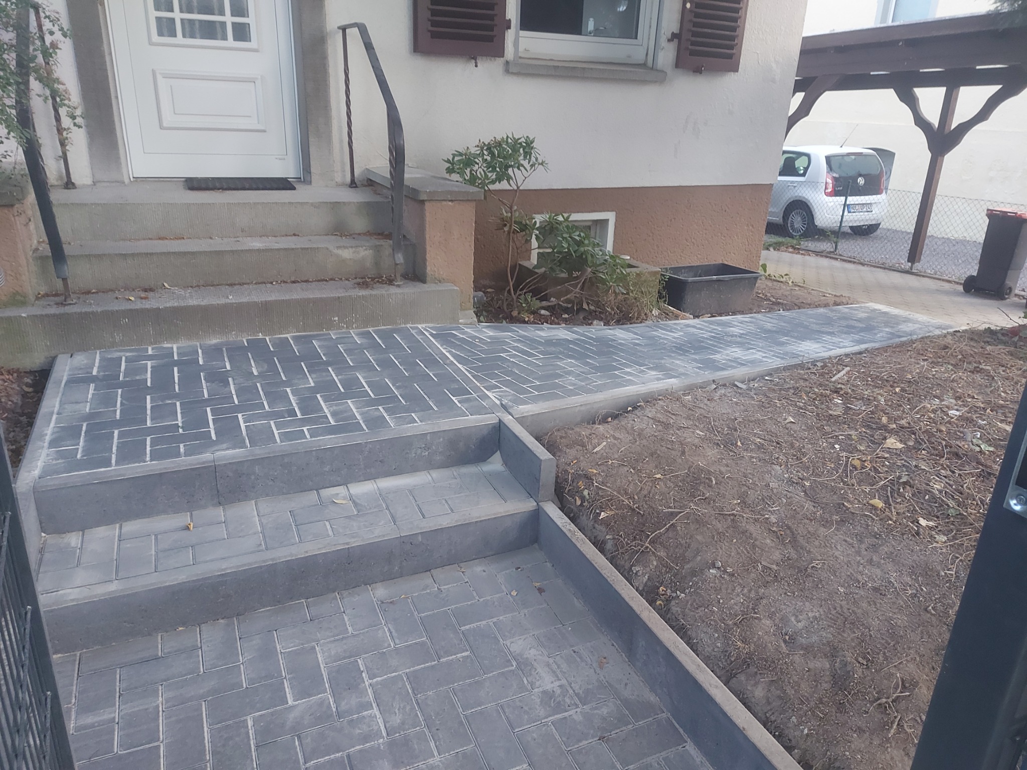 Ułożona szara kostka brukowa prowadząca do schodów wejściowych domu jednorodzinnego, widoczny fragment rabaty i podjazdu.