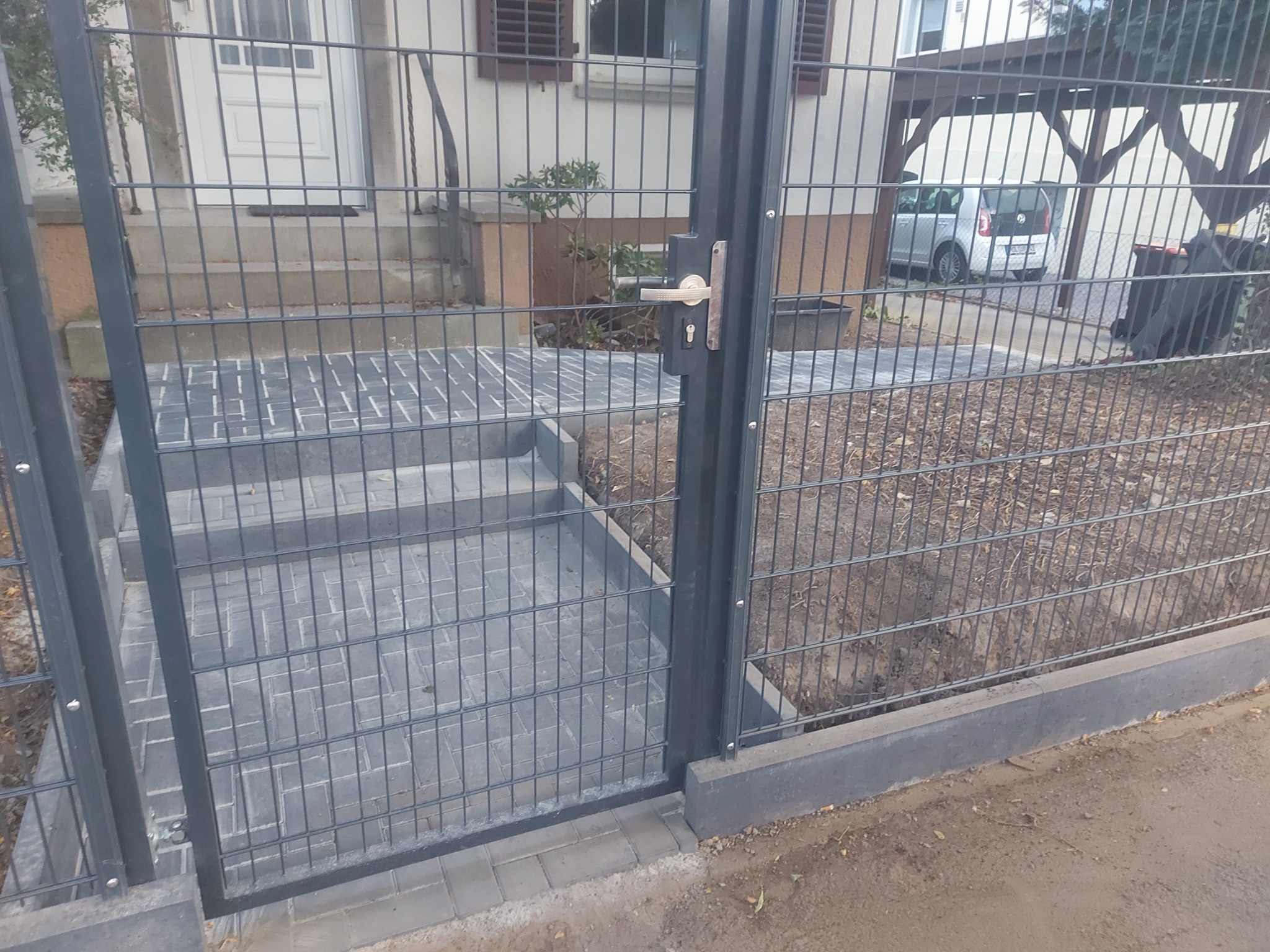 Nowoczesne ogrodzenie panelowe z bramką wejściową, szara kostka brukowa na schodach i podjeździe, widok na dom i zaparkowany samochód w tle.