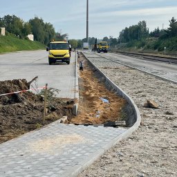 ML Pro Group Łukasz Marszał - Fragment układanej szarej kostki brukowej, widoczne żółte samochody dostawcze na tle torów kolejowych i ziemi. Prace w toku, widoczne materiały budowlane.
