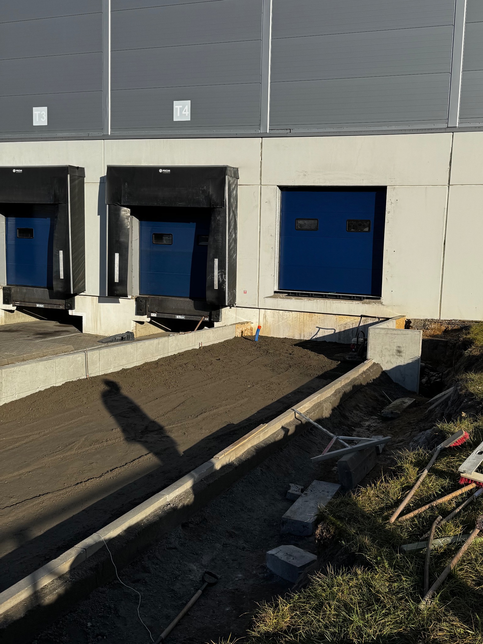 Przygotowanie terenu pod budowę rampy rozładunkowej, widoczne narzędzia i materiały budowlane, grunt wyrównany. Trzy bramy magazynowe.