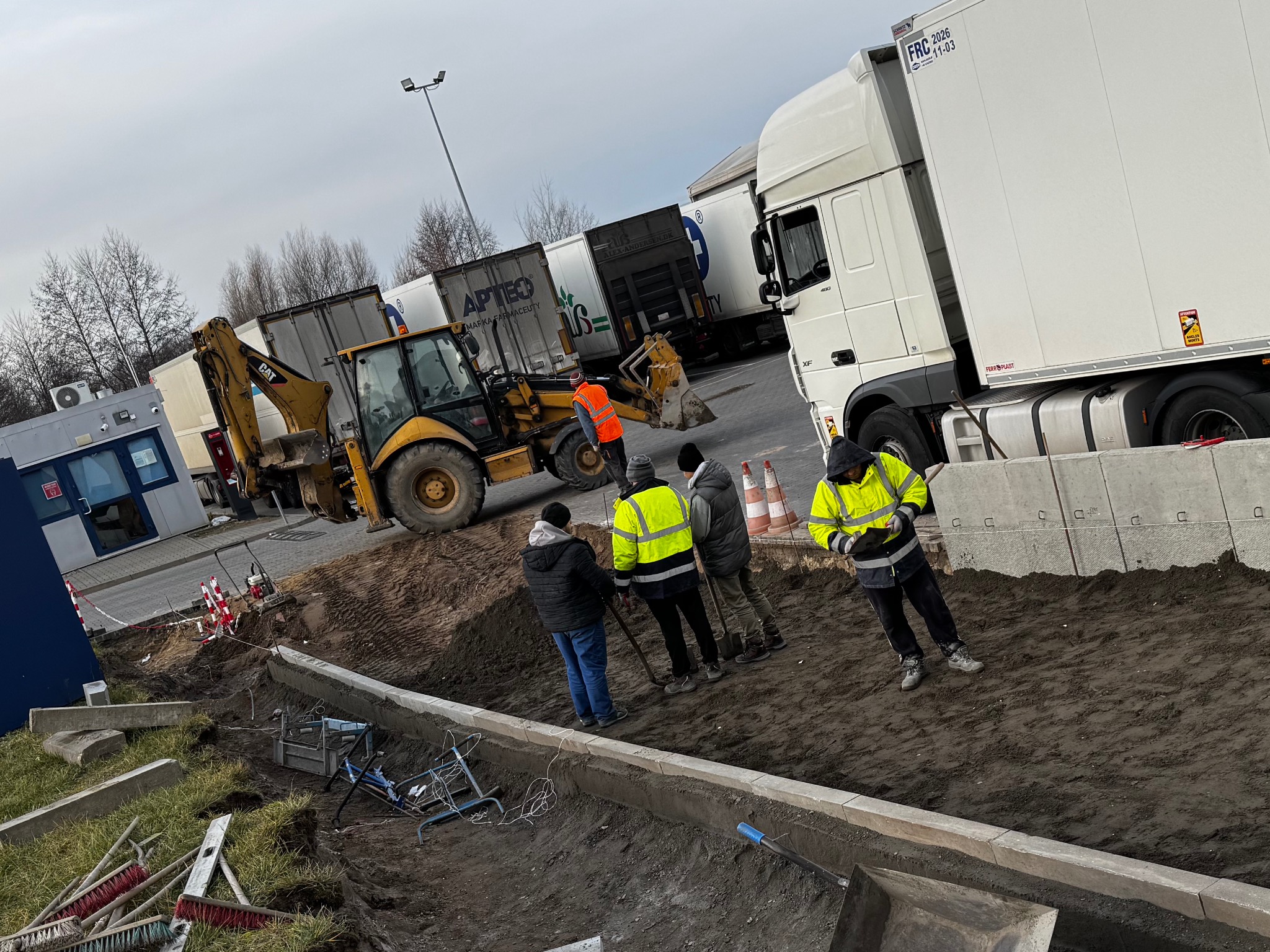 Prace ziemne z użyciem koparko-ładowarki i montaż obrzeży chodnikowych. Pracownicy w odblaskowych kamizelkach. W tle zaparkowane TIR-y.