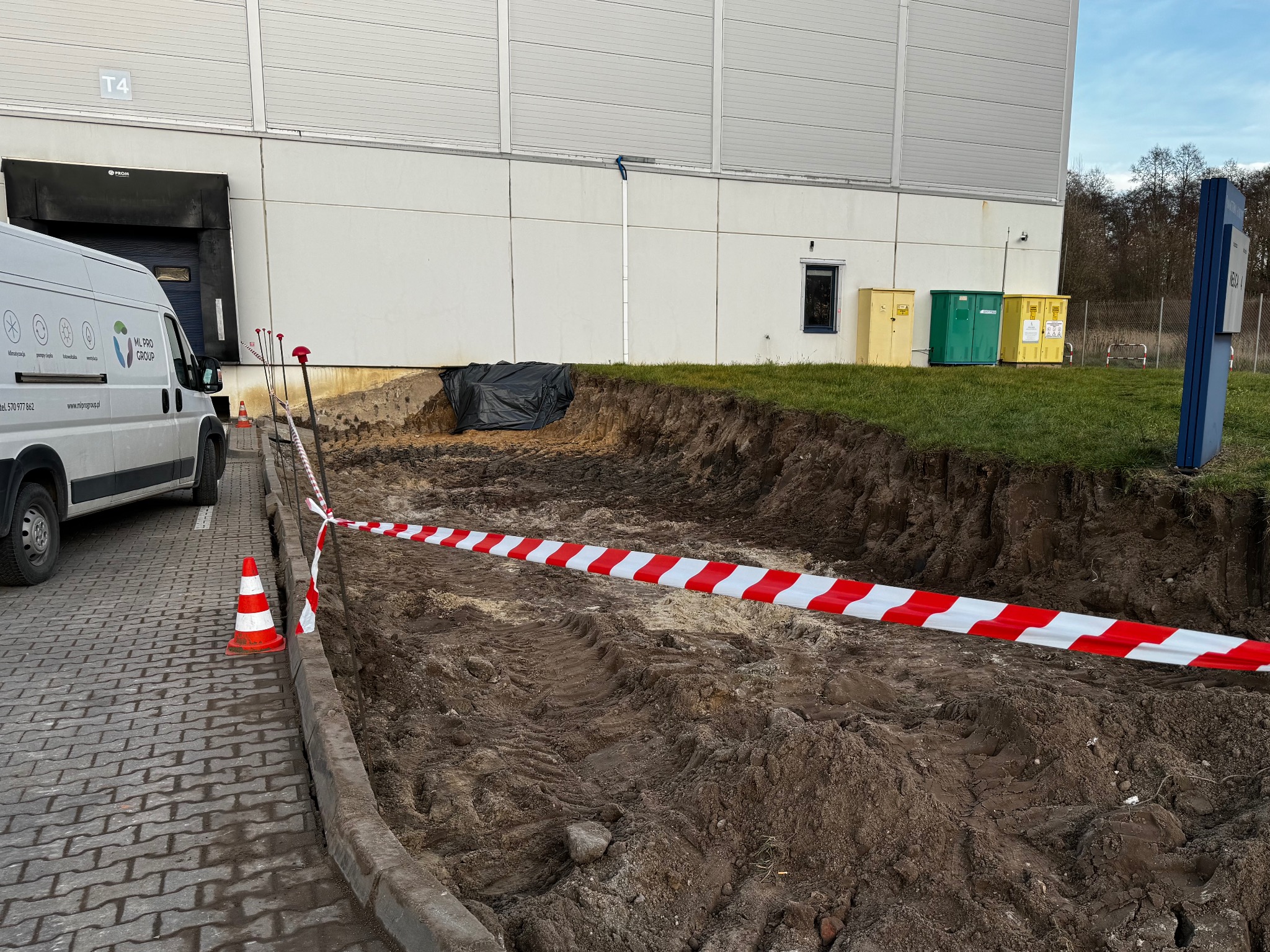 Wykonane wykopy ziemne przy budynku przemysłowym, zabezpieczone taśmą ostrzegawczą. Na pierwszym planie kostka brukowa i zaparkowany bus firmy ML PRO GROUP.