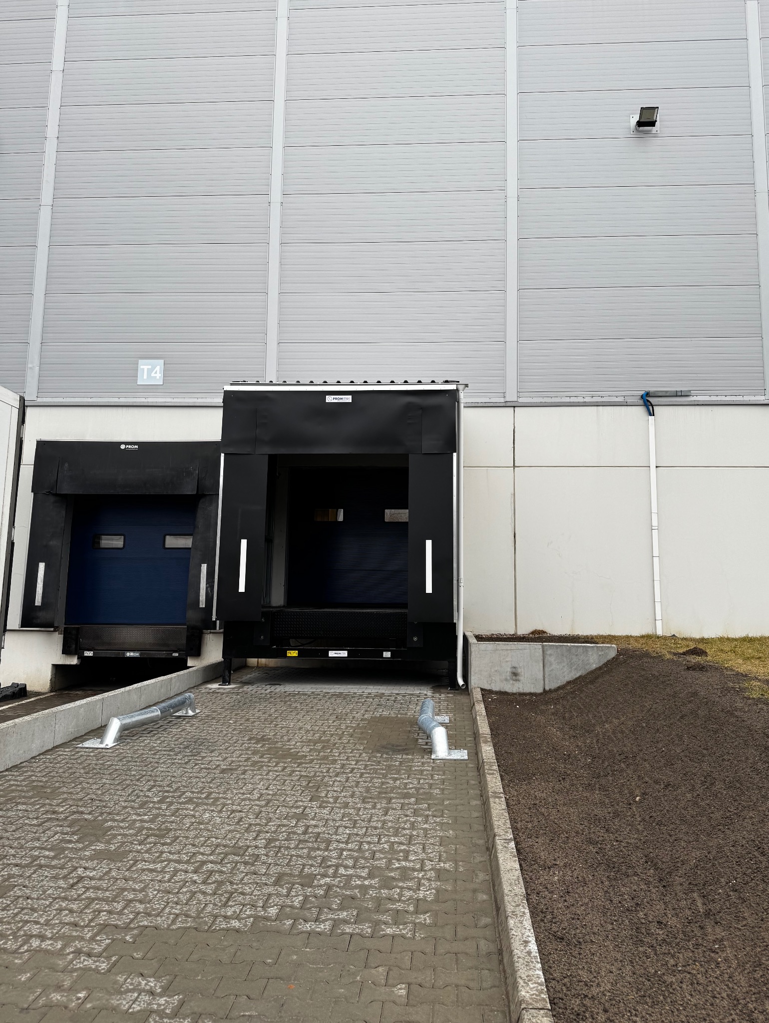Rampa przeładunkowa z kostki brukowej przed magazynem, widoczne czarne uszczelnienia dokowe i fragment ściany budynku z oznaczeniem T4.